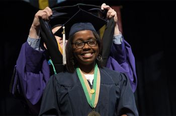 A GW Health Sciences student wearing graduation regalia receives a sash from a faculty member